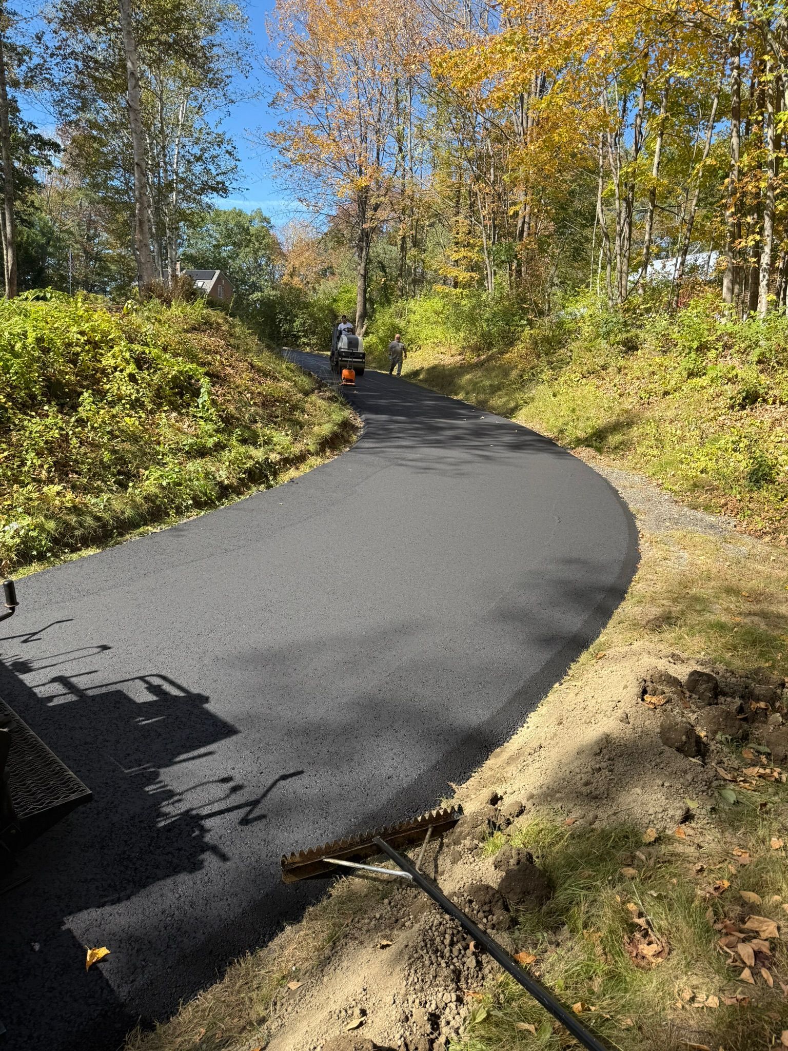 A freshly paved black asphalt driveway winds through a wooded area with autumn foliage under a clear blue sky.