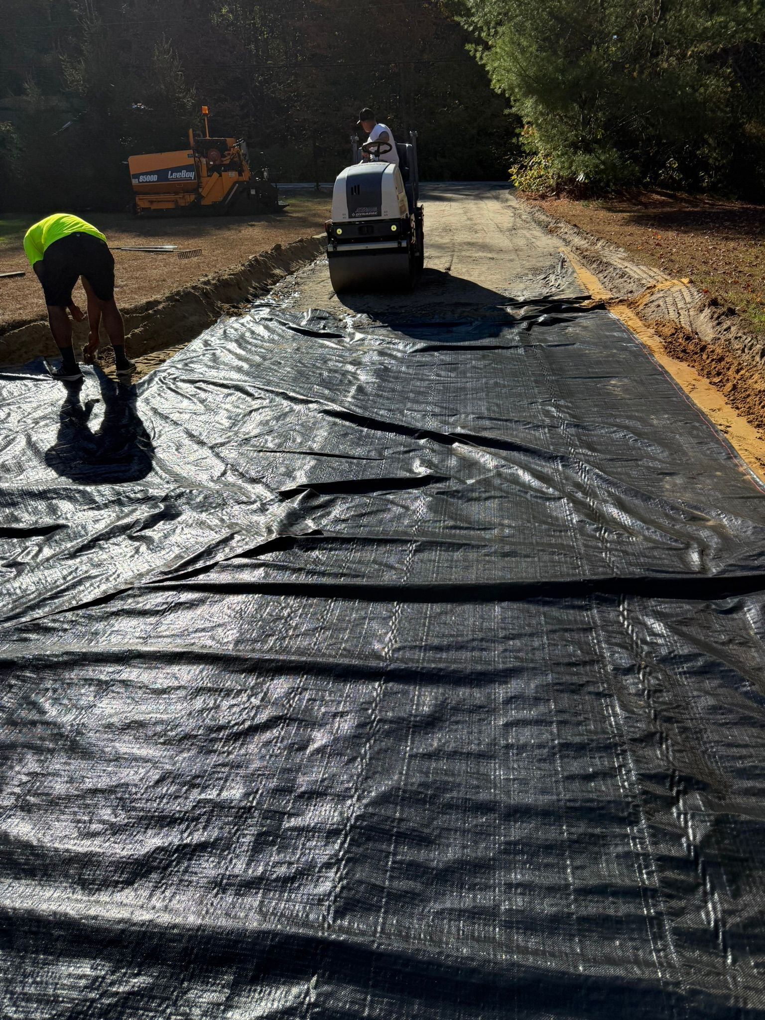 A person in a bright yellow shirt stands near a road construction site where a roller flattens black landscape fabric.