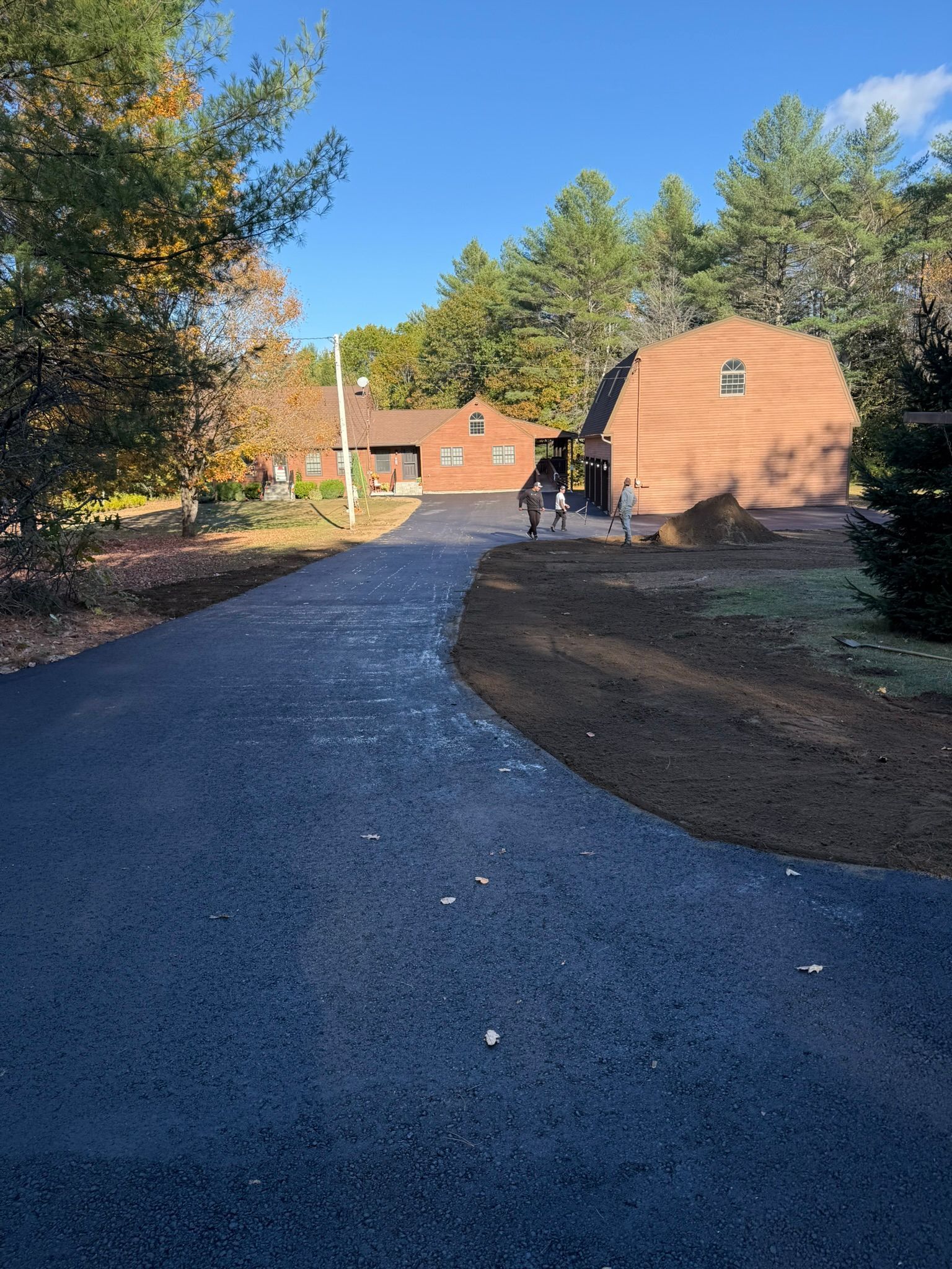 A freshly paved asphalt driveway leading toward a house and a large brown barn-style building on a sunny day.