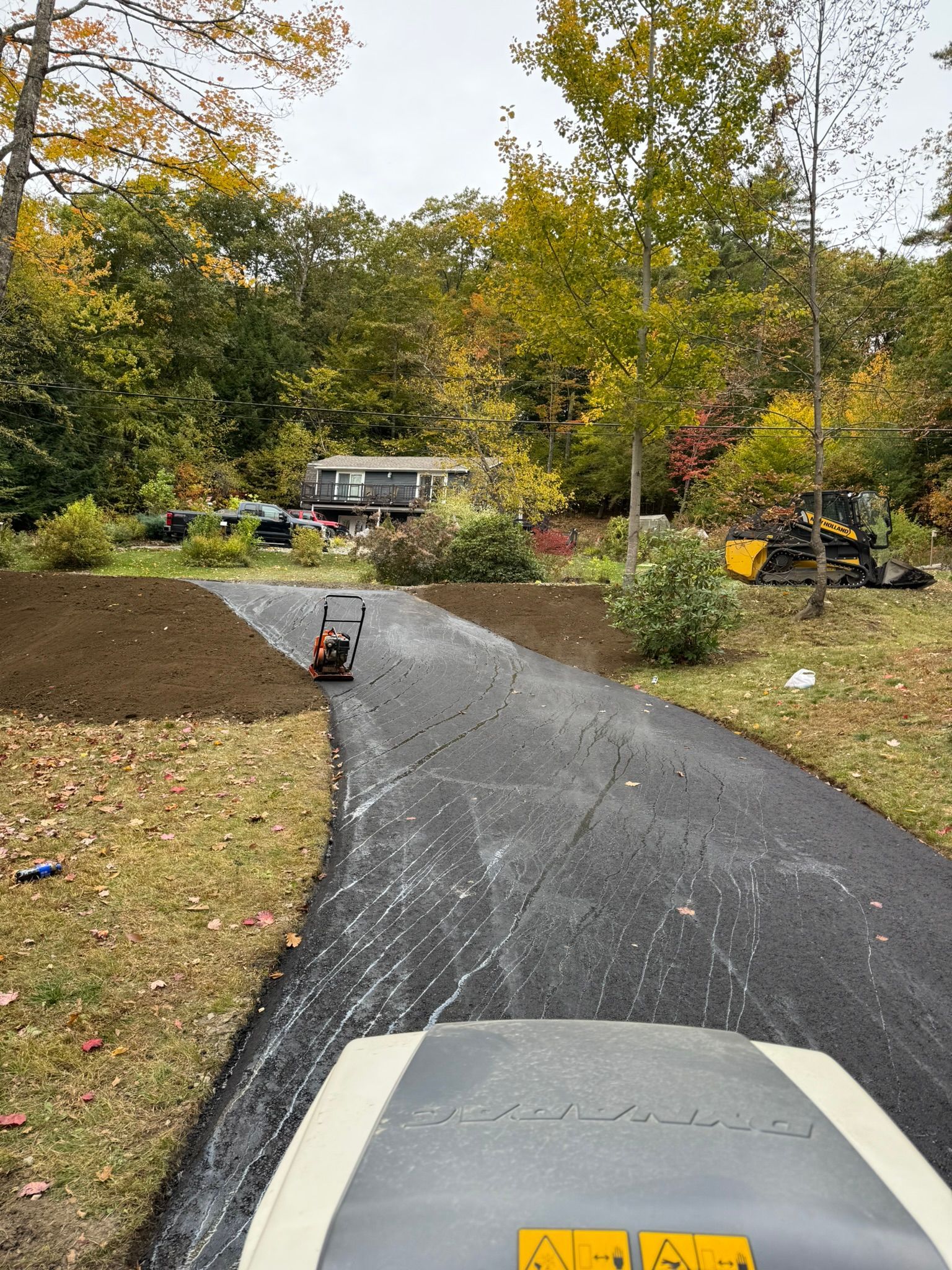 A view from a construction vehicle looking down a freshly paved asphalt driveway toward a house in an autumn setting.