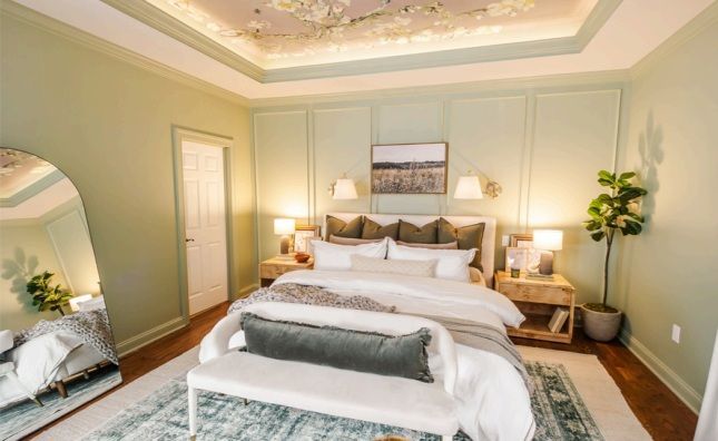 A bedroom with light green walls and a floral ceiling. A bed with white and gray bedding sits in front of a paneled wall. A full-length mirror and a potted plant are visible.