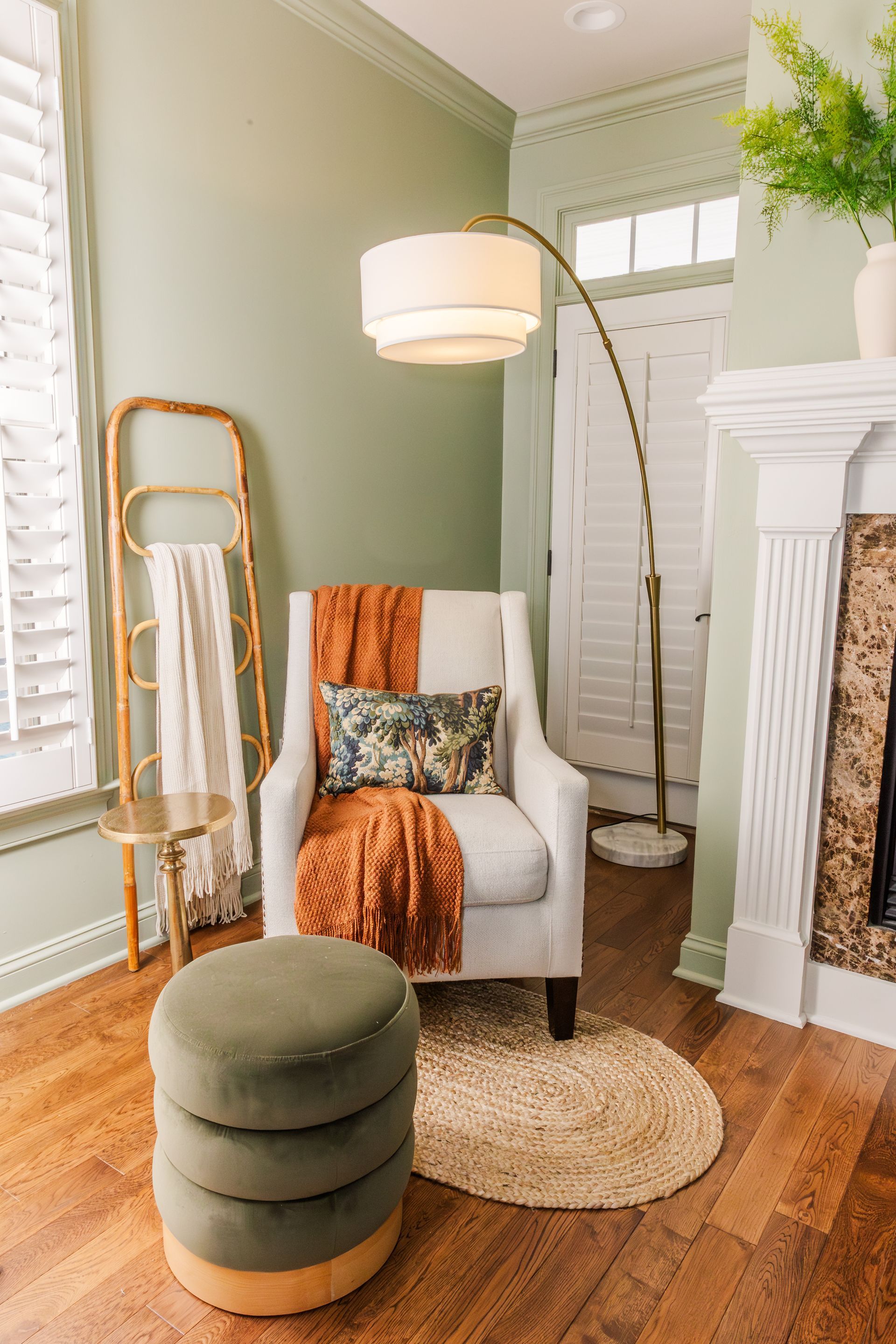 Cozy reading nook with a white armchair, orange throw, and arched floor lamp; set against a sage green wall, and wooden floor.