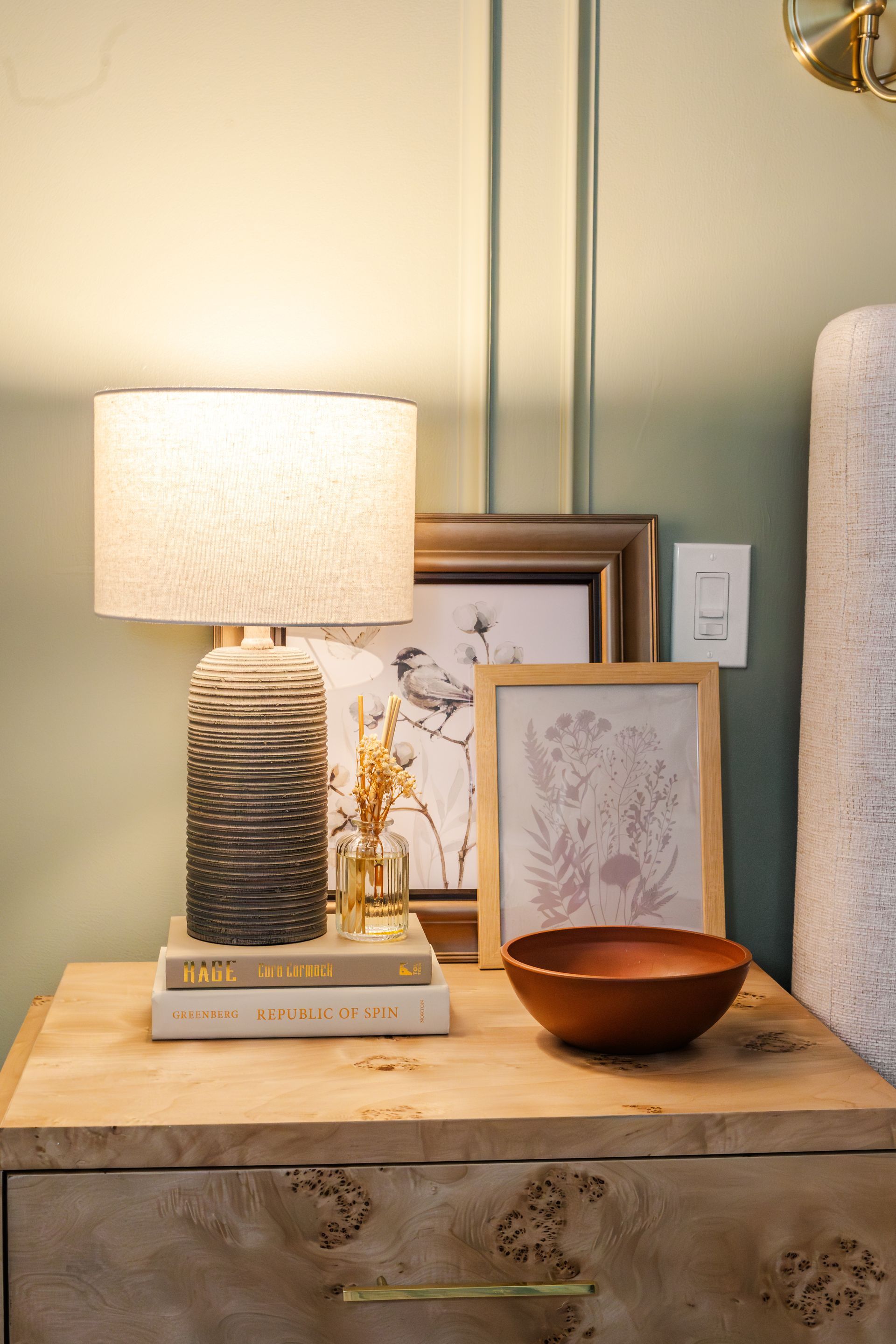 Nightstand with lamp, books, framed art, and wooden bowl, in a room with green walls.