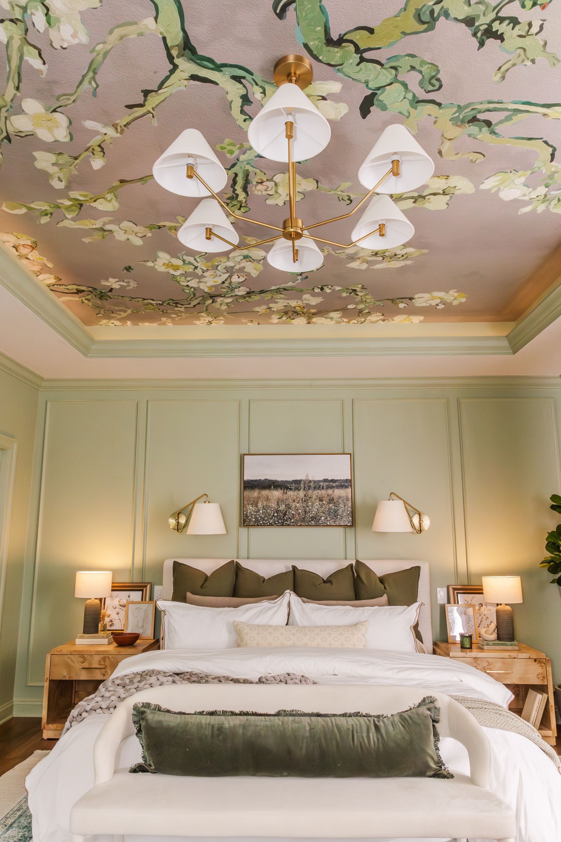 A bedroom with a sage green wall, a decorated ceiling, and a large bed with various pillows and a bench.