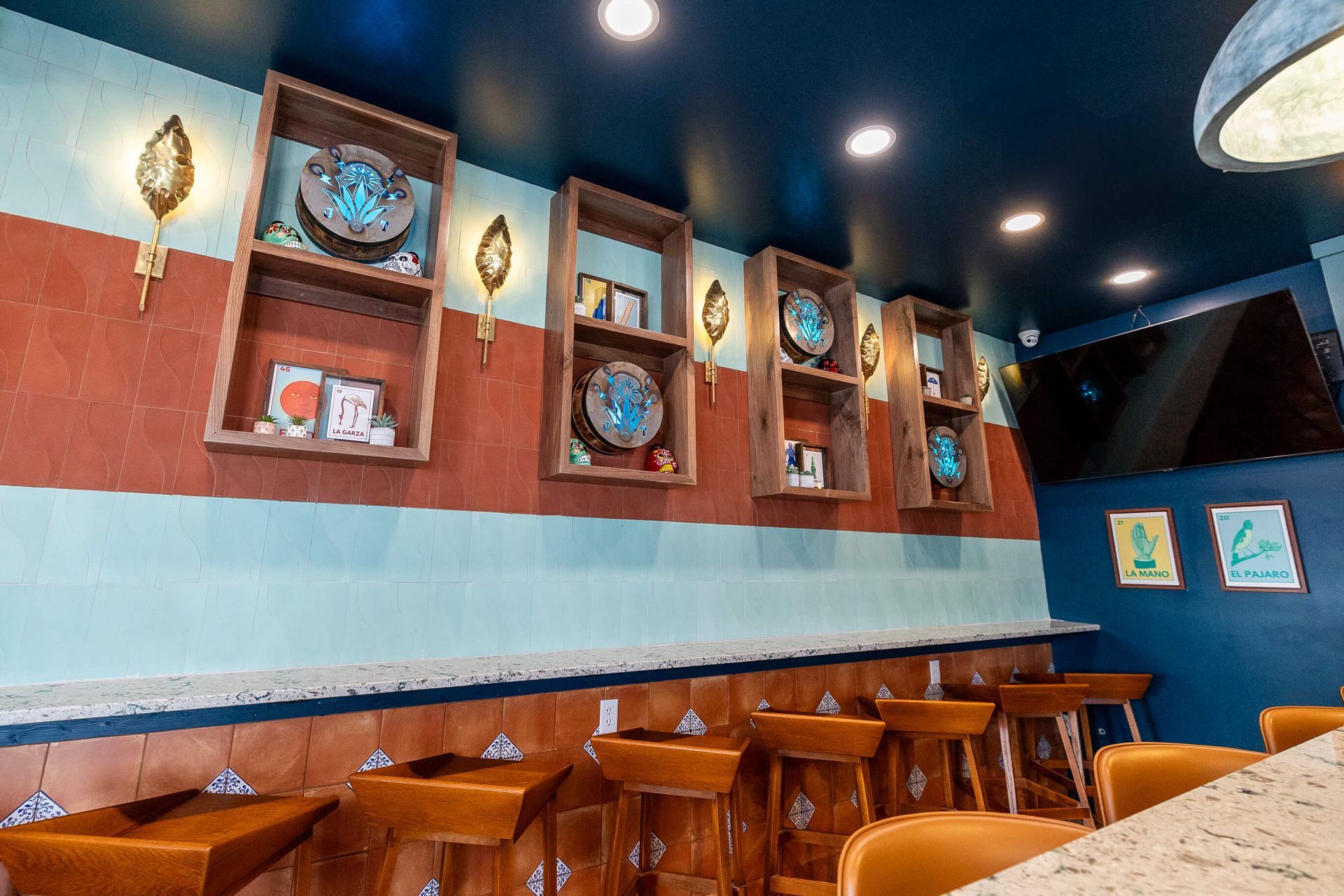 Interior of a restaurant with a bar, wood shelving, and colorful tile and walls. Barstools are at the counter.