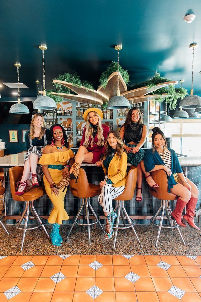 Seven people seated at a bar, striking poses. Diverse outfits, vibrant colors. Interior with plants, blue walls, orange floor.