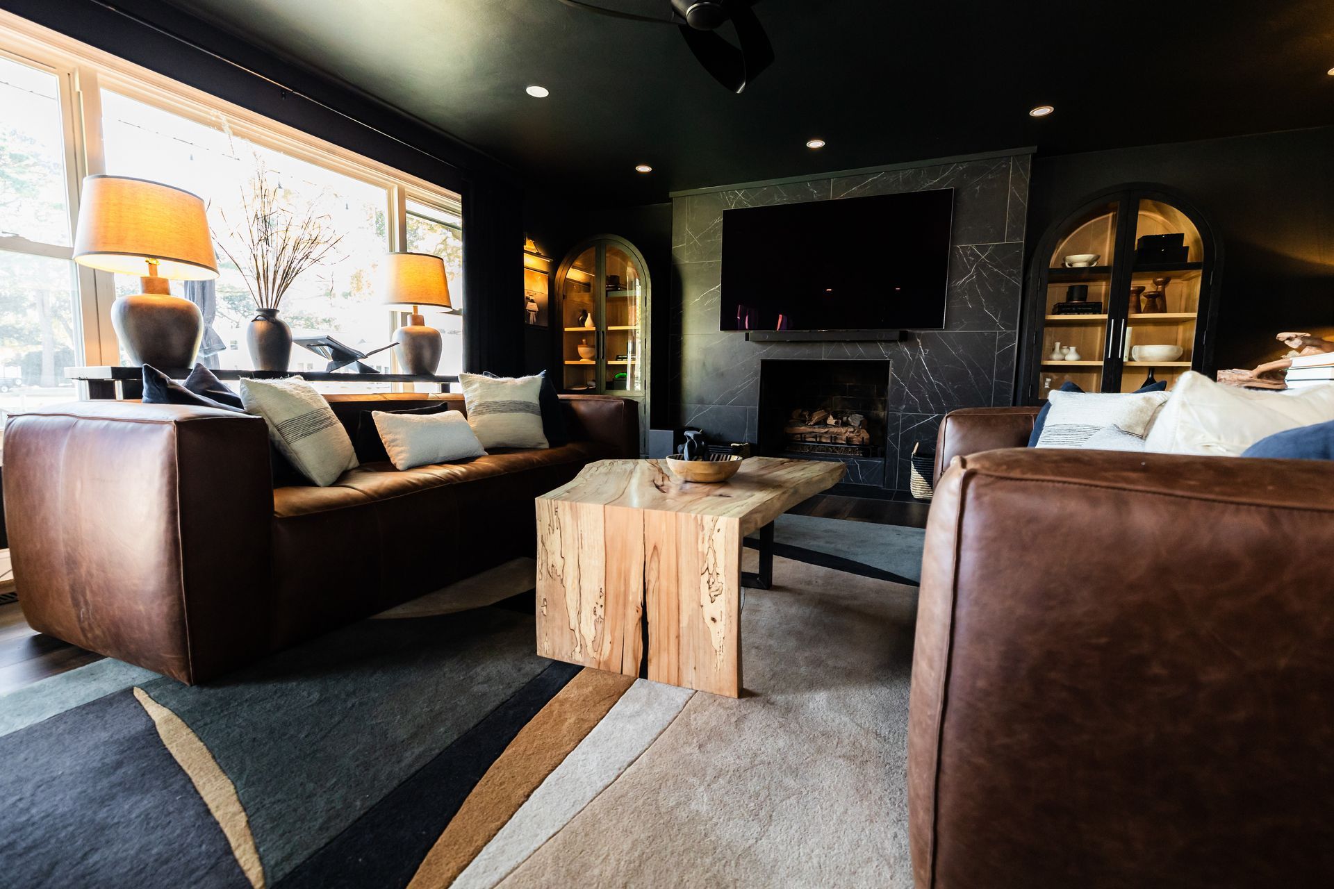 A dark, modern living room with leather couches, a wooden coffee table, a fireplace, and a large TV.