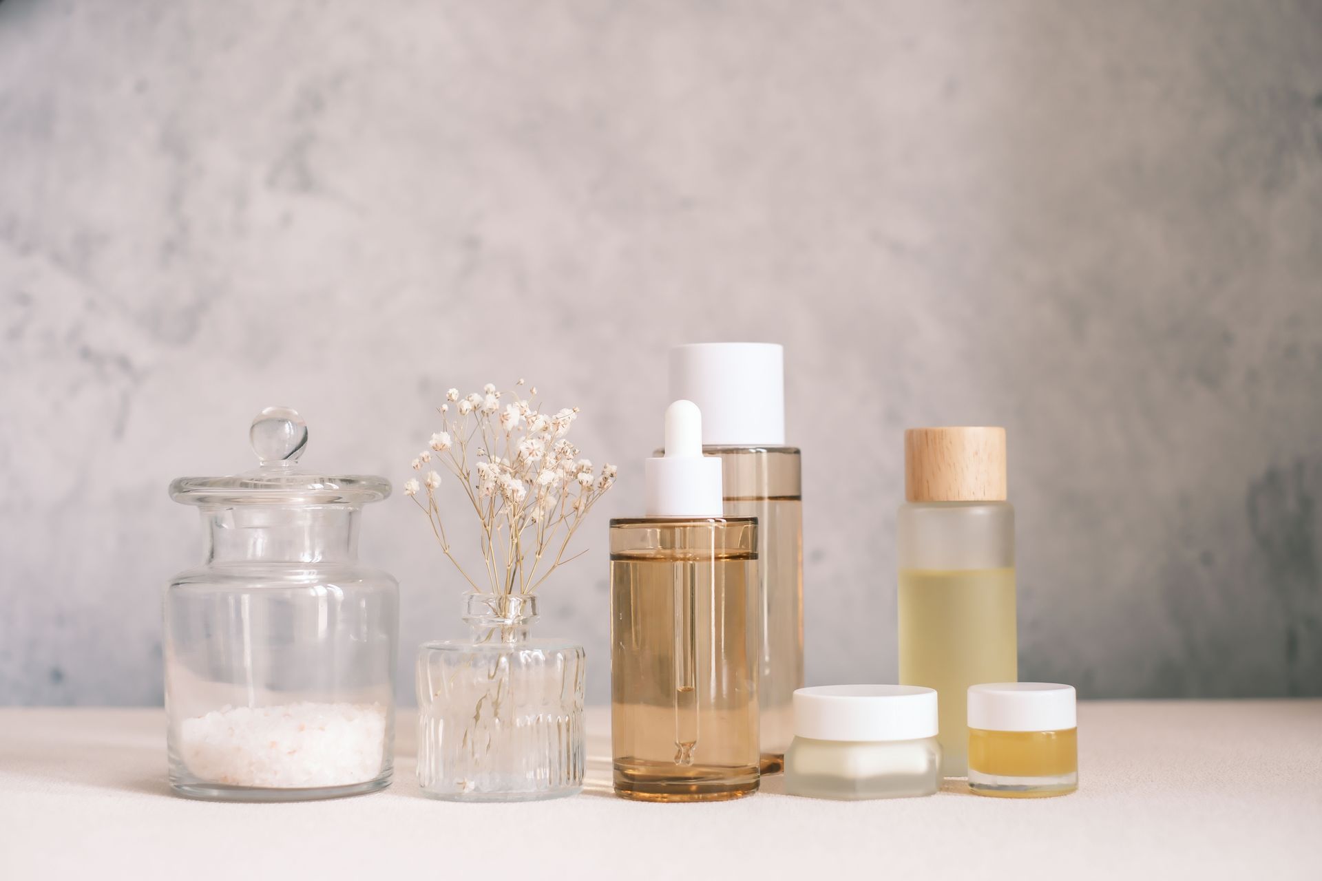 A collection of skincare products including bottles, jars, and a vase with white flowers on a table against a gray wall.