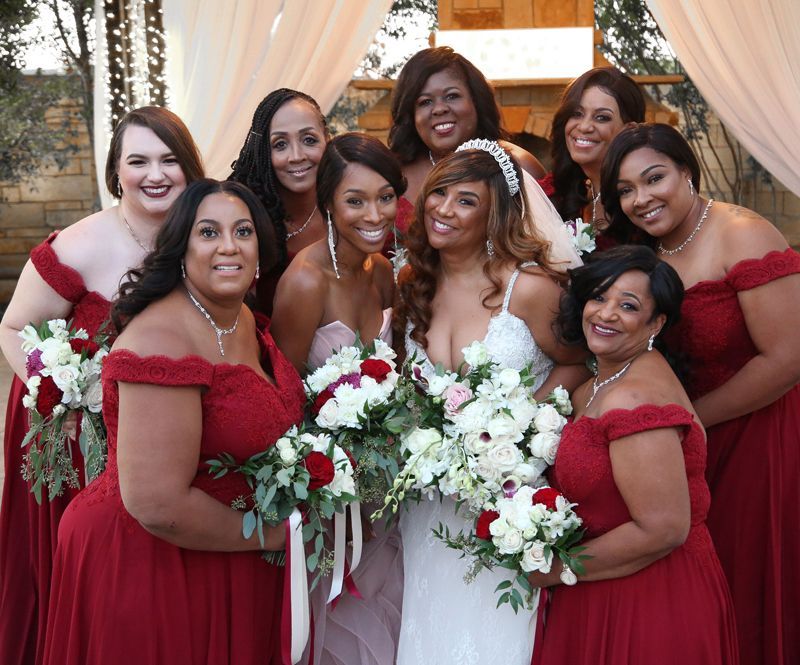 Bridal party in red dresses posing with the bride in a white gown and bouquets under a draped arch
