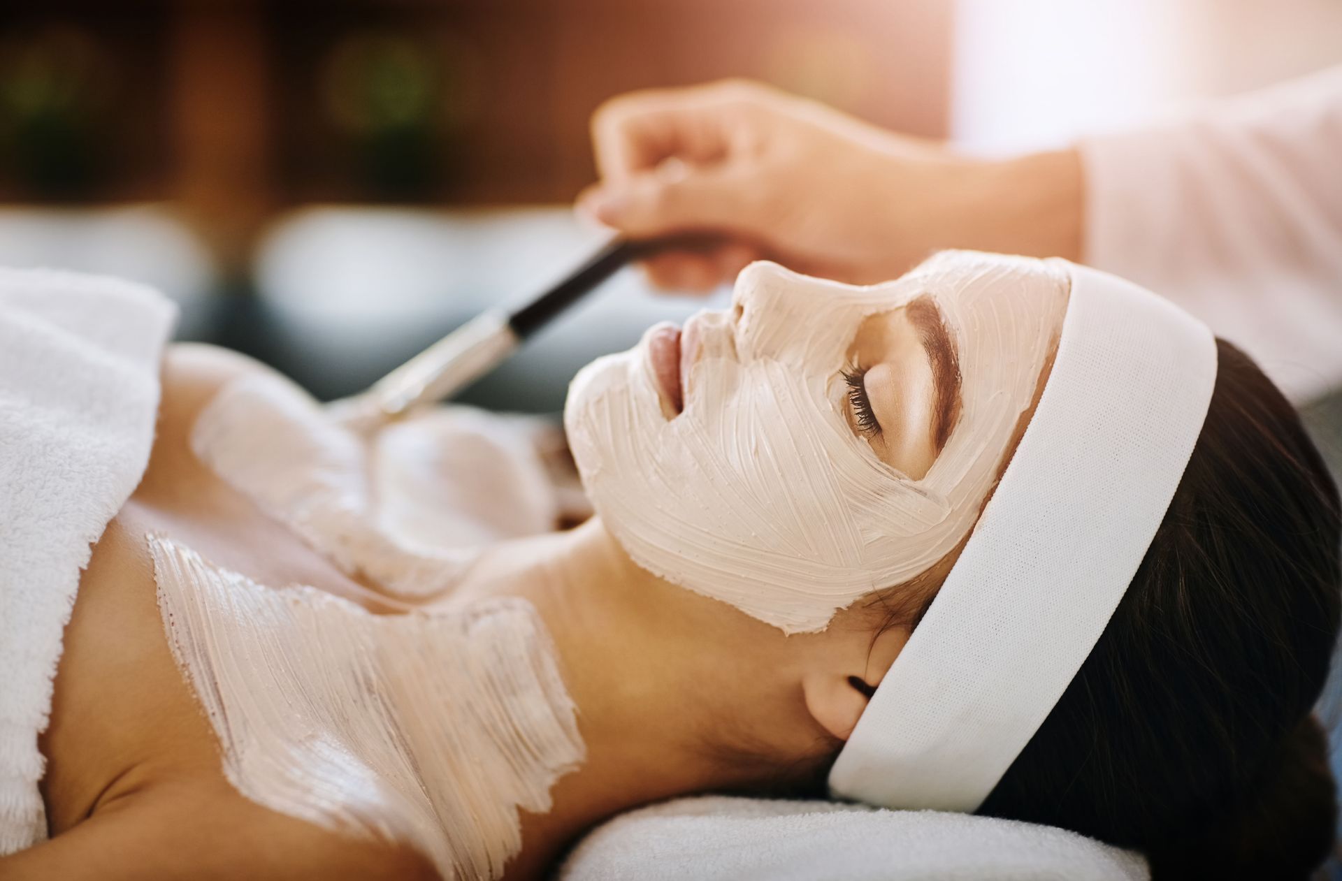 A professional applying a light-colored face mask to a person’s face and chest with a brush in a spa setting.