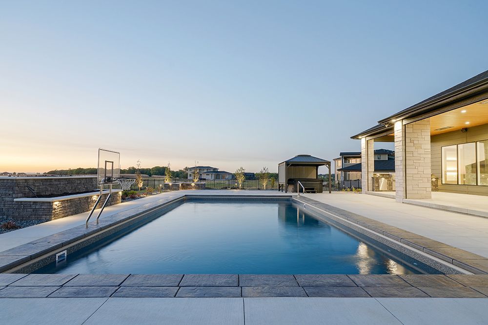 Rectangular pool at dusk beside a house with a basketball hoop. The sky is clear.