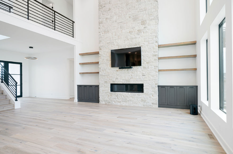 Spacious living room with stone fireplace, built-in shelves, and a TV; wood floors and large windows.
