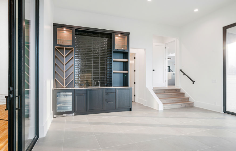 Dark gray built-in bar with wine storage, cabinets, and a sink, in an empty room with stairs in the background.