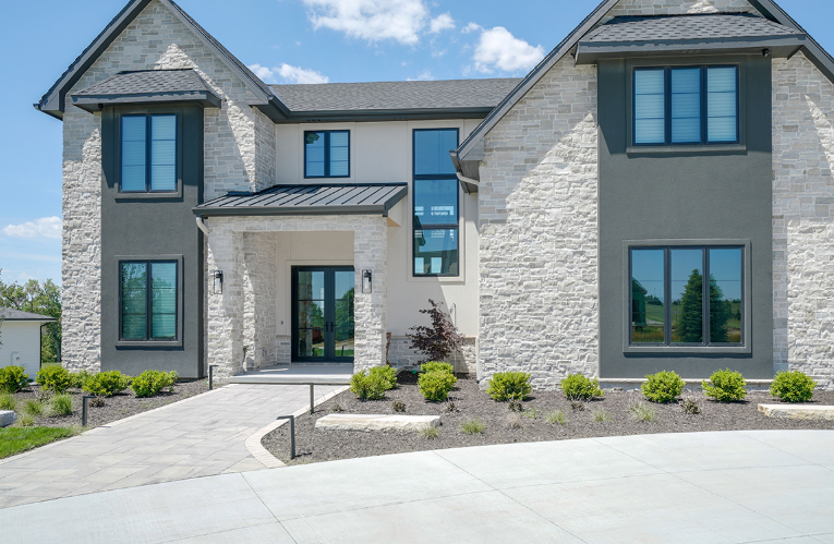 A light stone and grey stucco house with a driveway and bushes, under a blue sky.