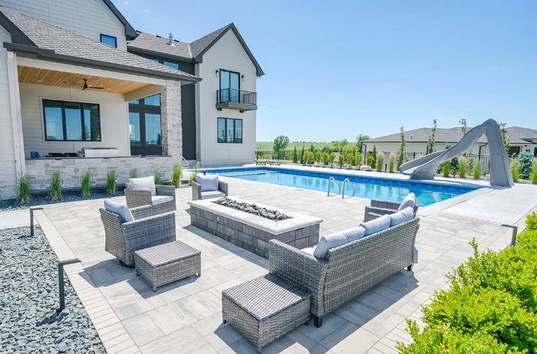 Backyard patio with wicker furniture, a rectangular pool, and a gray pool slide under a blue sky.