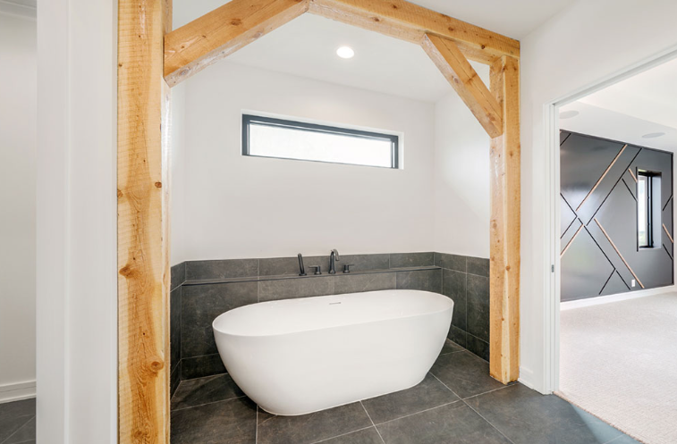 Freestanding white bathtub under wooden beam arch in modern bathroom.