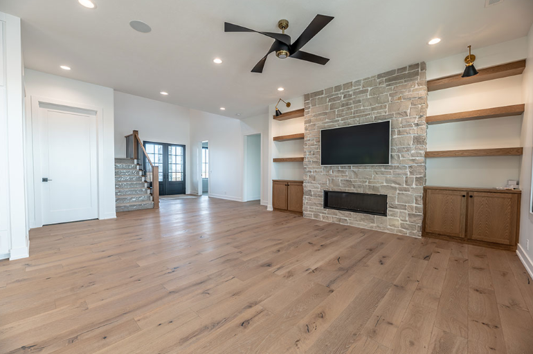 Empty, bright living room with hardwood floors, a stone fireplace, built-in shelves, and a ceiling fan.