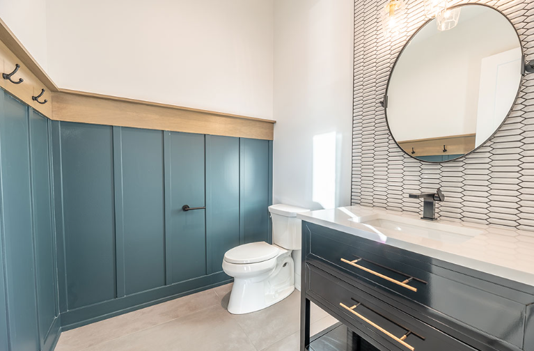 Blue-paneled bathroom with a toilet, black vanity, round mirror, and patterned backsplash.