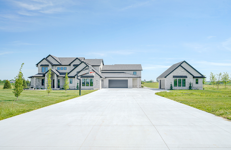 Large gray and white house with long driveway and detached garage, under a blue sky.