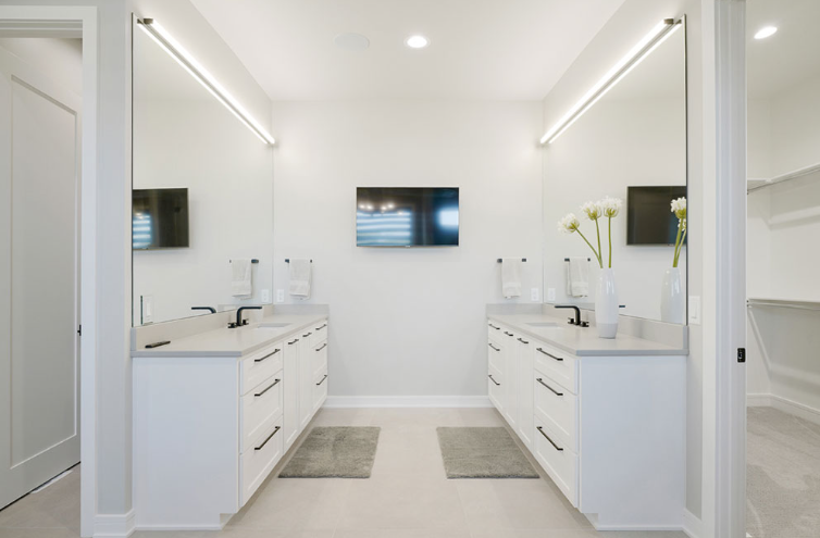 Modern white bathroom with two sinks, large mirrors, and a TV mounted on the wall.