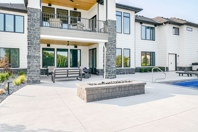 Backyard patio with fire pit, pool, and modern white house with stone columns.