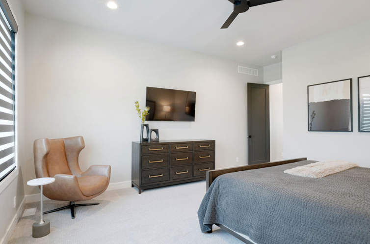 Modern bedroom with a chair, dresser, bed, and television. Neutral colors with light walls and a dark brown door.