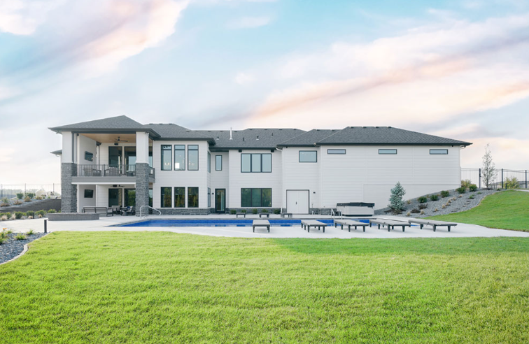Large white house with a pool, patio, and green lawn under a partly cloudy sky.