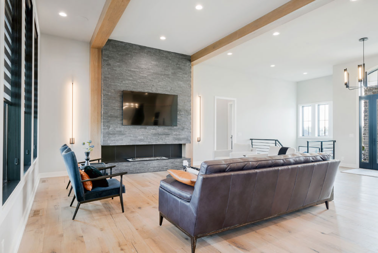 Spacious modern living room with fireplace, gray stone wall, wood beams, leather couch, and blue chairs.