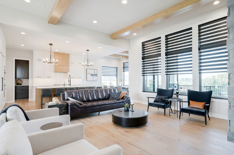 Spacious modern living room with light wood floors, large windows, dark furniture, and open kitchen.