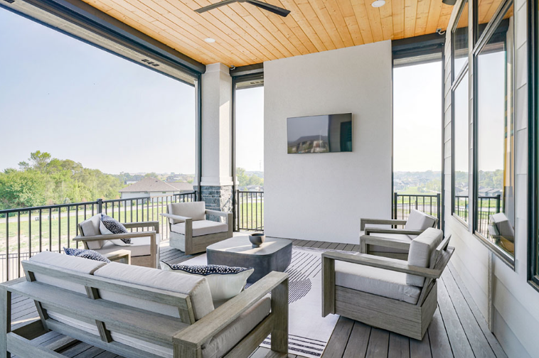 Outdoor patio with seating area, tv, and wood ceiling.  Black railing and view of trees.