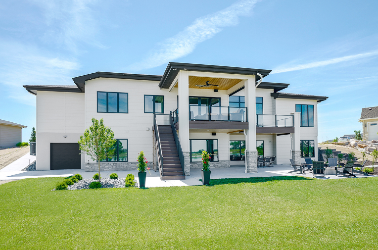 Rear view of a modern white house with a deck, stairs, and manicured lawn under a blue sky.