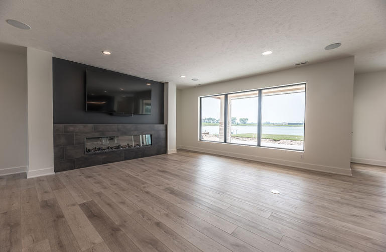 Modern living room with fireplace, TV, and large windows overlooking a body of water. Light wood floors.
