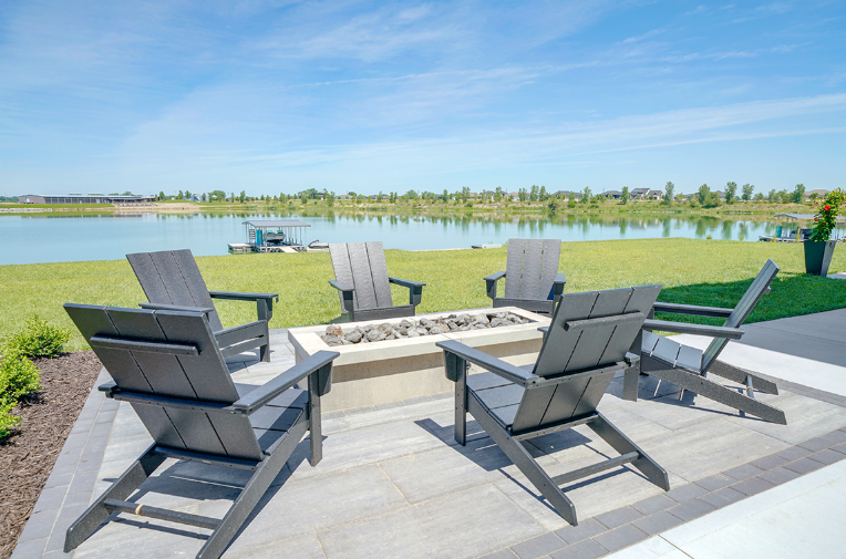 Patio with gray chairs around a fire pit, overlooking a lake and green lawn under a blue sky.