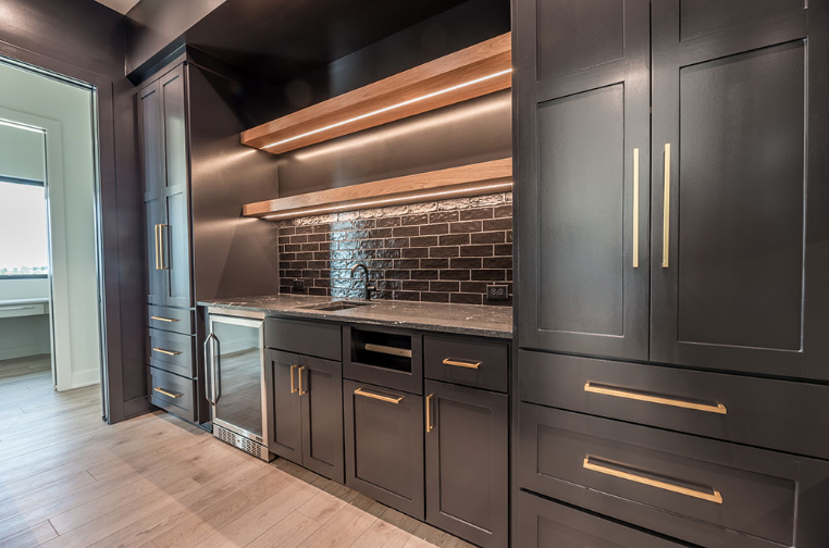 Dark gray built-in bar with gold hardware, brick backsplash, and wooden shelves with under-cabinet lighting.