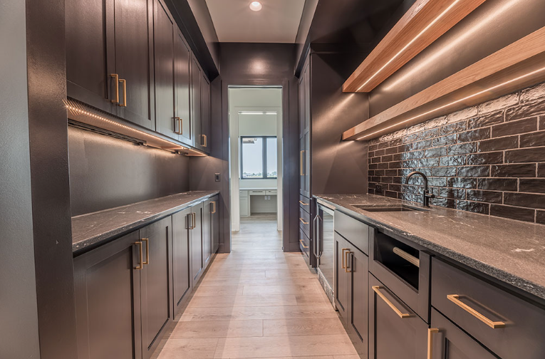 Dark gray built-in pantry with light countertops, wood shelves, and recessed lighting, leading to a small desk area.