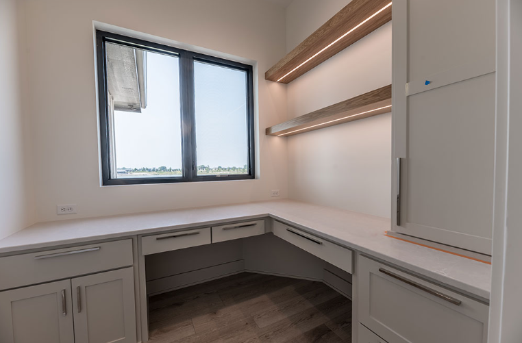 Home office with white built-in desk, cabinets, and floating shelves. Window on the left.