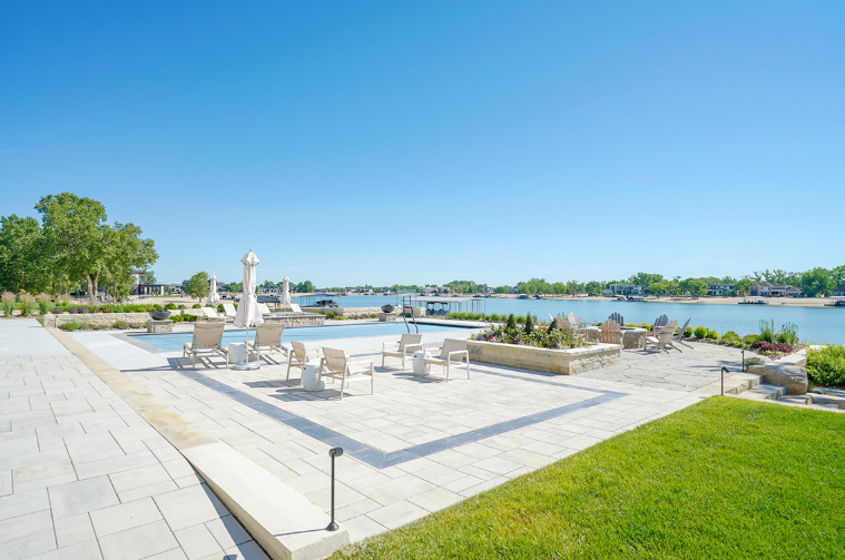 Poolside patio overlooking a waterway with lounge chairs, blue sky, and green grass.