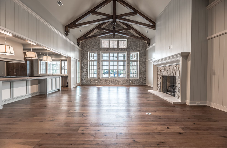 Spacious room with wood floor, stone fireplace, and large windows. Exposed wooden beams on high ceiling.