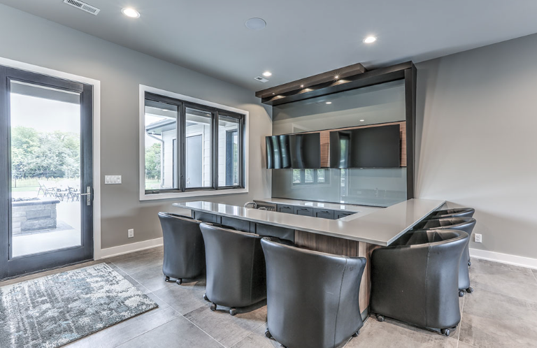 A modern bar area with gray walls, leather seating, countertop, and multiple TVs. Doorway to a patio.