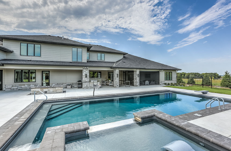 Large house with a rectangular pool and a spa. Blue sky and green field in the background.
