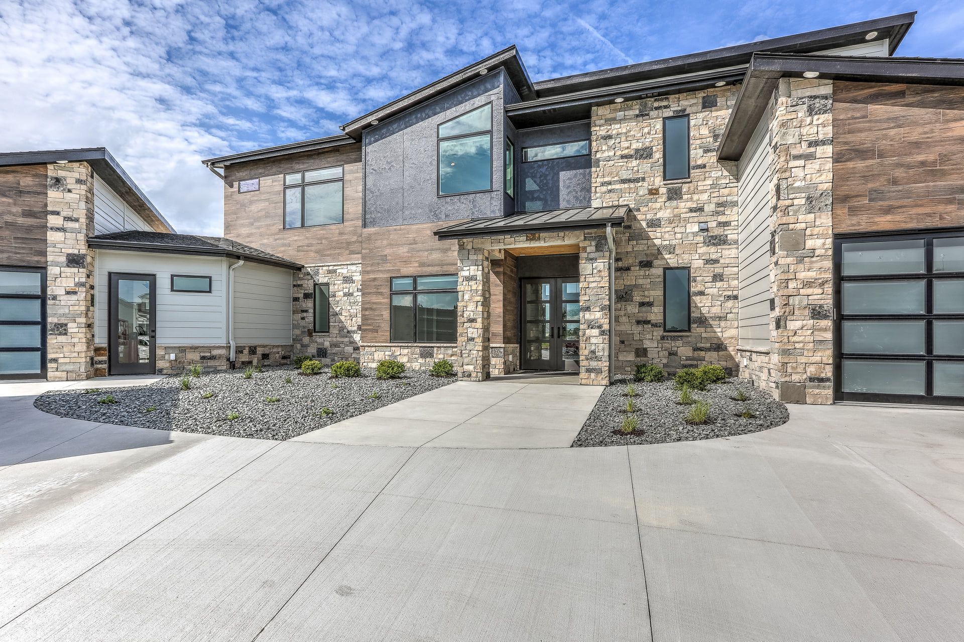 Modern two-story house with stone and brick exterior, glass garage doors, concrete driveway, and rock landscaping.