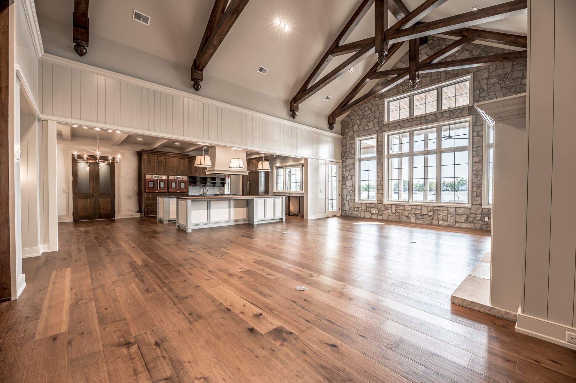 Spacious, open-concept living area with wooden floors, exposed beams, stone wall, large windows, and kitchen.
