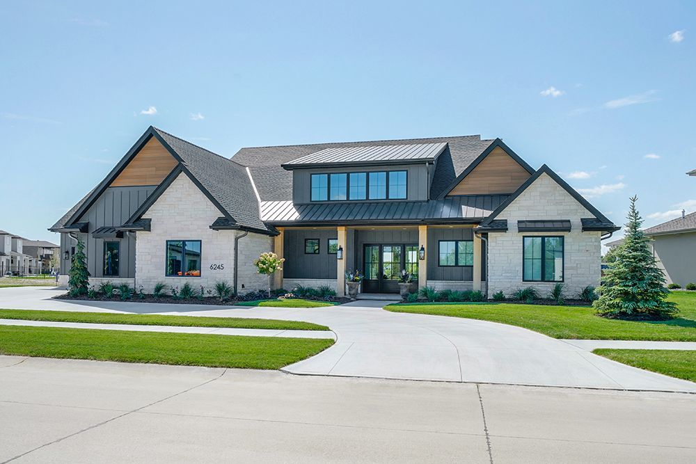 Modern, two-story house with gray and white brick facade, wooden accents, and a long driveway on a sunny day.