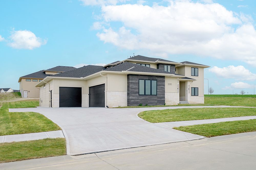 Modern two-story house with a two-car garage, beige siding, dark trim, and a concrete driveway. Blue sky with clouds.