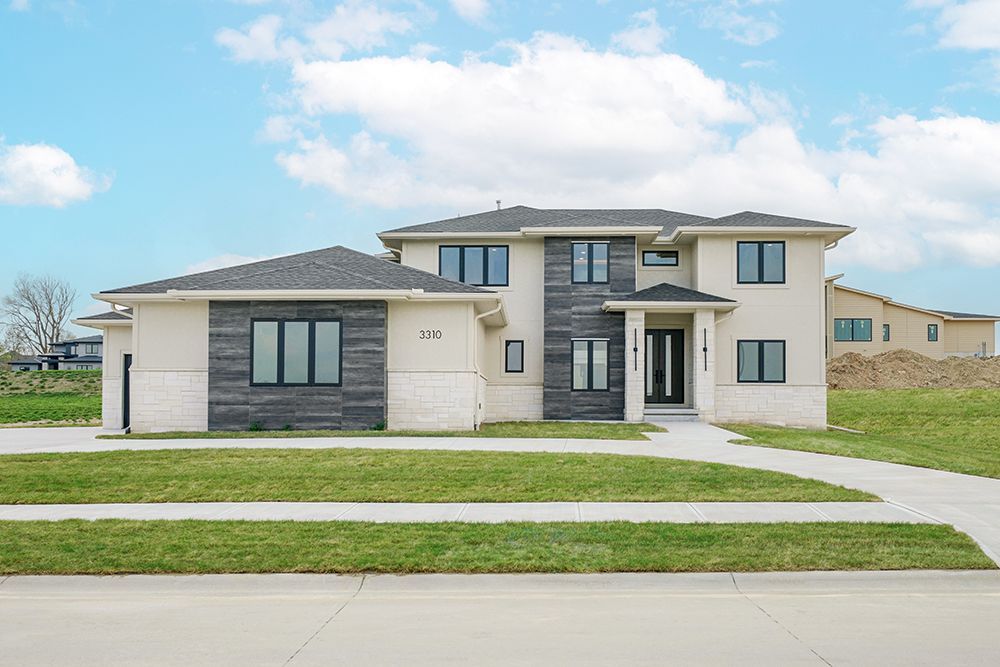 Two-story modern house with light stucco and gray stone accents, black-framed windows, and a paved walkway.