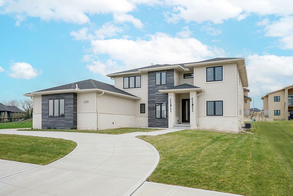 Modern two-story house with light stucco exterior, dark window frames, and curving driveway under a cloudy sky.