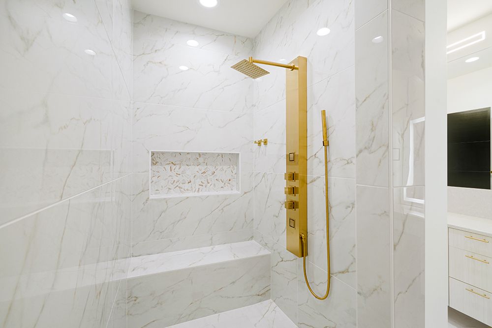 White marble-tiled shower with gold fixtures, including a rain showerhead, built-in bench, and niche.