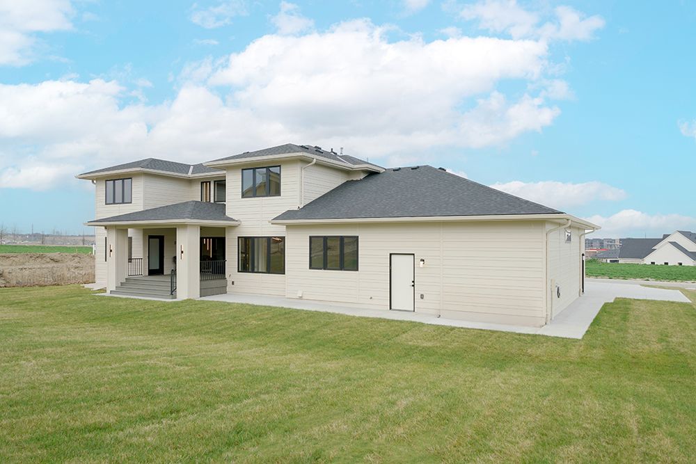 Modern, two-story beige house with dark roof on a green lawn under a cloudy blue sky.
