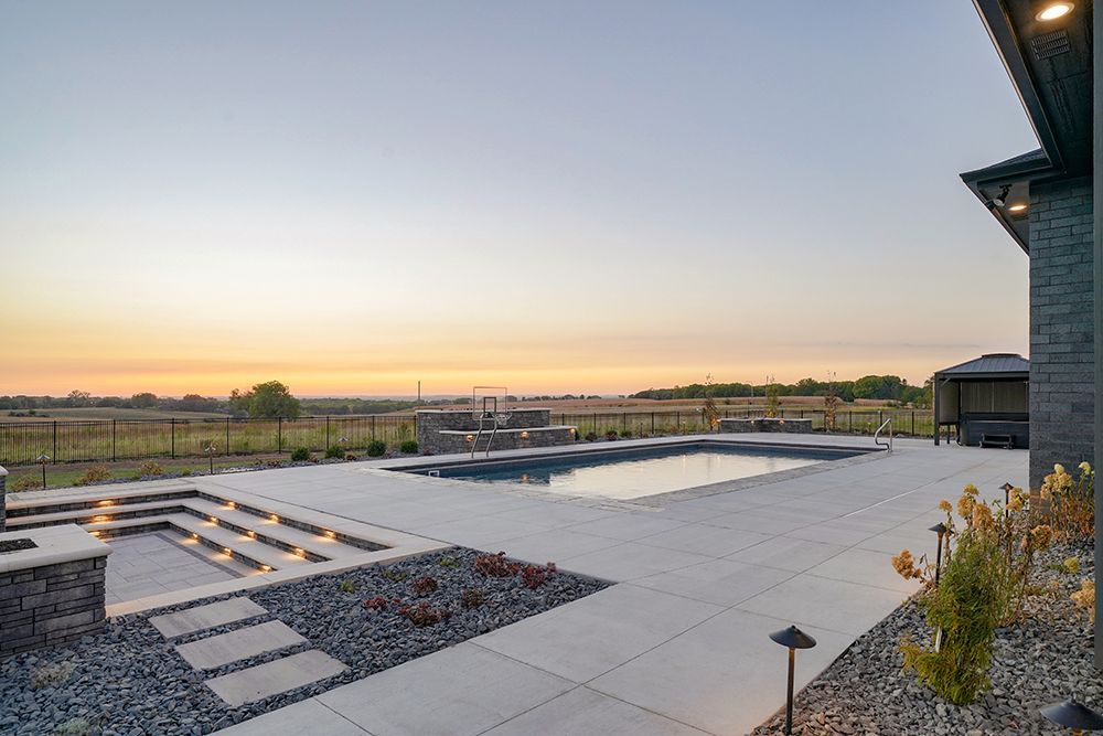 Patio with pool, steps, and distant view at sunset. Stone, concrete, and landscaping.