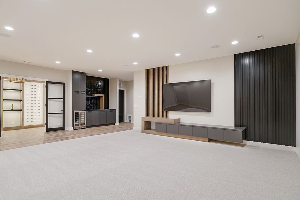 Modern, empty basement with light gray carpet, TV, and black panel wall. Includes cabinets and built-in bar.